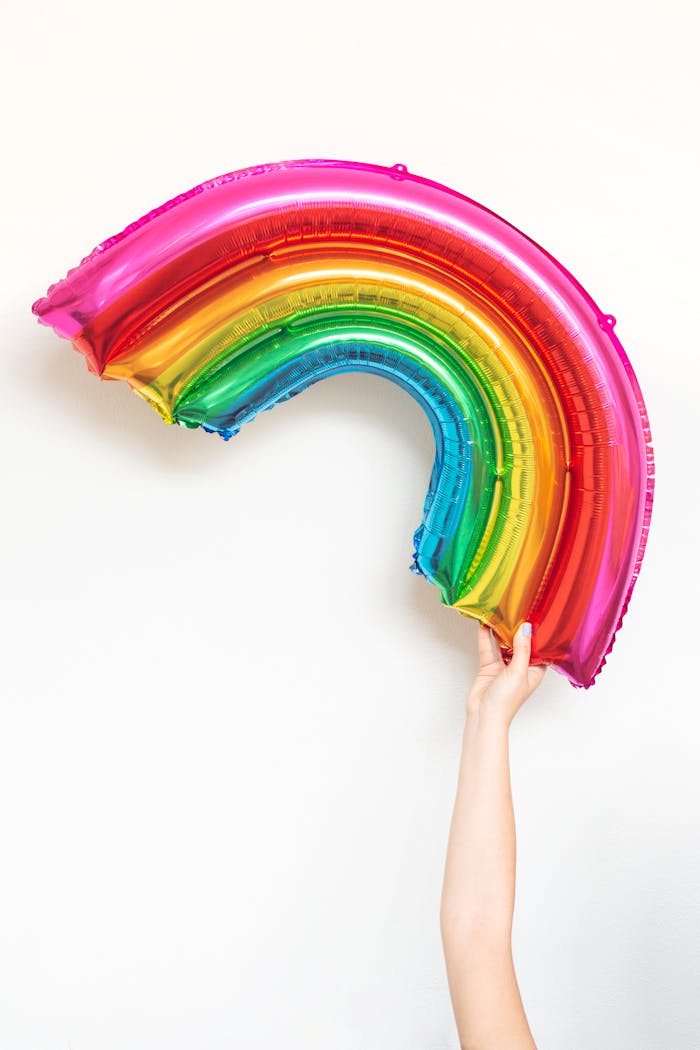 A bright rainbow balloon being held up by a hand against a white background.
