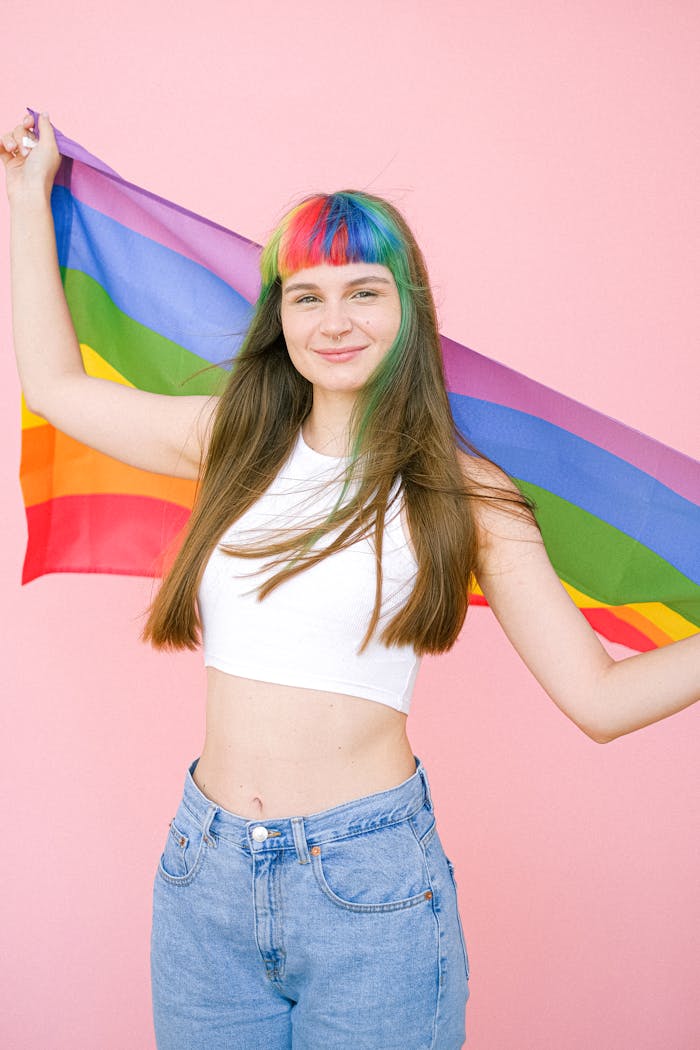 Joyful young woman with rainbow hair holding a pride flag in front of pink background.